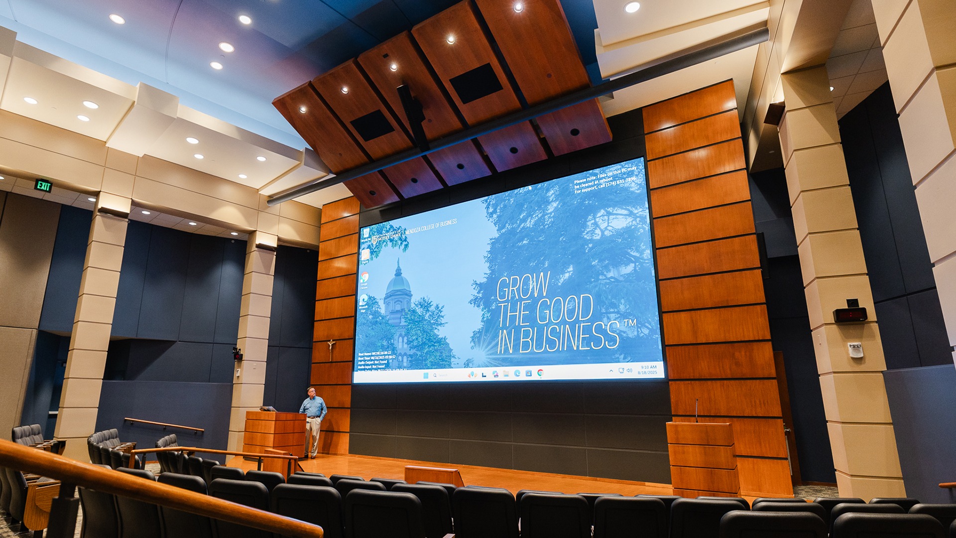 Large auditorium with projector screen down and man standing on stage